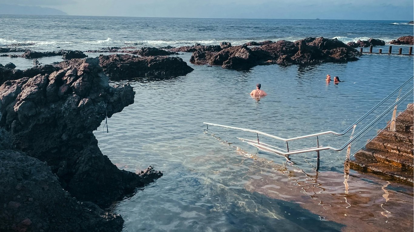 Piscina natural de agua de mar entre coladas de roca volcánica, con una escalera metálica y tres bañistas al fondo